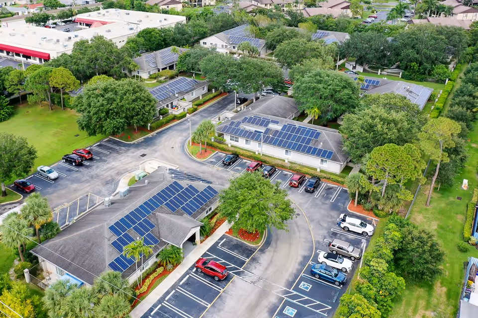 Aerial view of a senior living facility named Cottages at Palm Beach showing multiple buildings with solar panels on the roofs, surrounded by trees and greenery. There are parking lots with several cars parked, and well-maintained landscaping throughout the property.