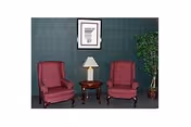 Two red upholstered armchairs with wooden legs are positioned on either side of a small round wooden table with a lamp on it. A framed picture hangs on the dark green wall behind the chairs, and a tall potted plant is placed to the right of the chairs.