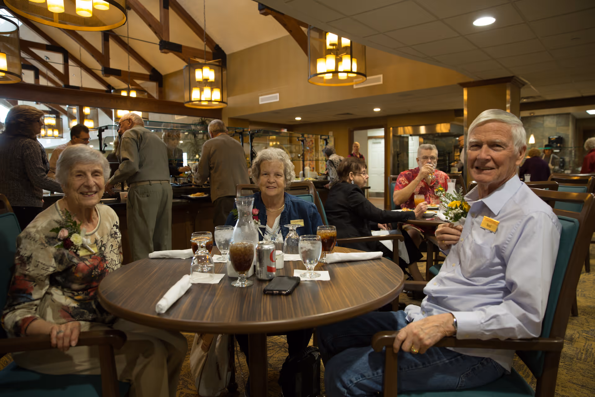 Three smiling elderly residents seated at a round table in a busy retirement community dining room.