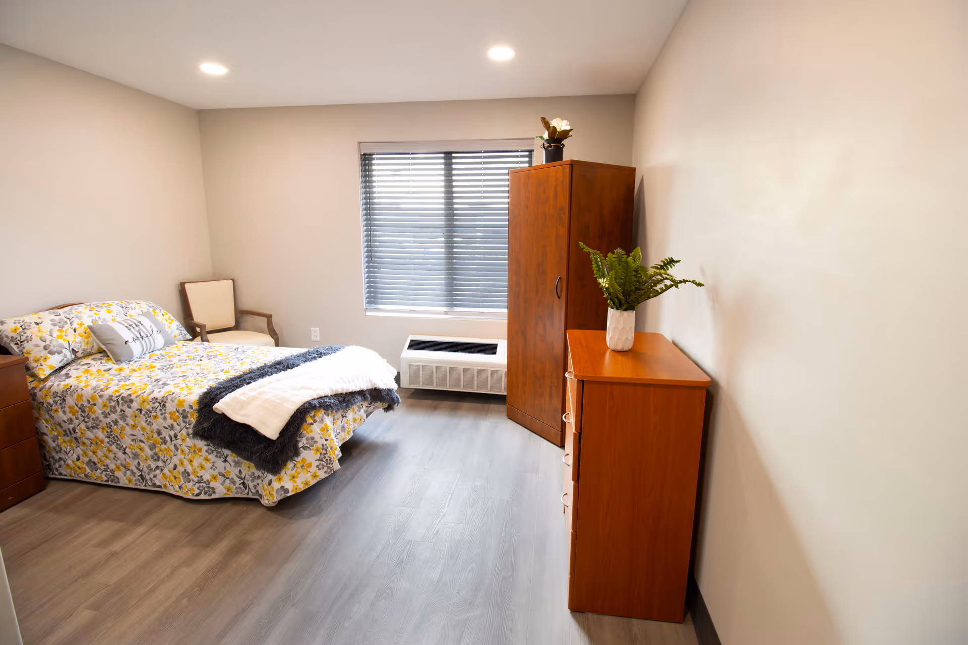 A bright and clean bedroom with a bed covered in floral bedding, a wooden dresser with a potted plant, a wooden wardrobe, a chair in the corner, and a window with blinds letting in natural light.