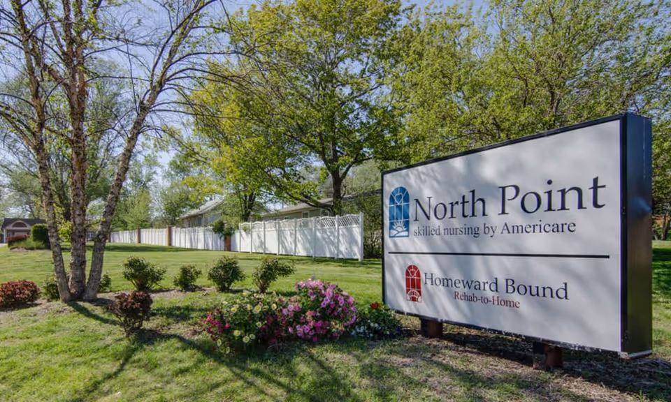 Outdoor view of the North Point skilled nursing facility sign surrounded by green grass, flowering bushes, and trees with a white fence and building in the background under a clear sky.