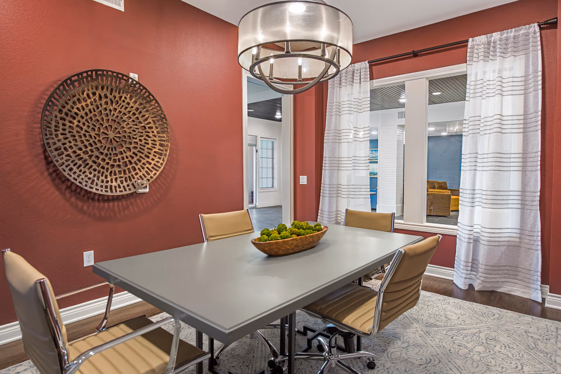 Modern dining room with a rectangular gray table, tan chairs, a large pendant light, and decorative wall art on a red wall.
