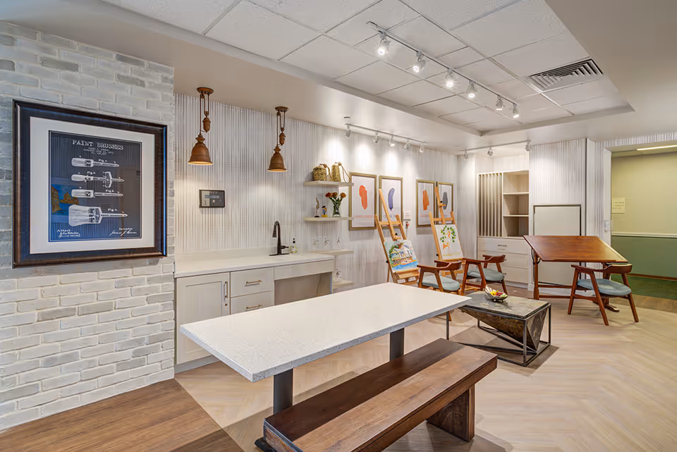A well-lit interior room with a long white table and wooden bench in the foreground. The back wall has a countertop with a sink, shelves with decorative items, and framed artwork. There are two wooden chairs with cushions and easels displaying paintings. The ceiling has track lighting and pendant lights, and the floor is light wood.