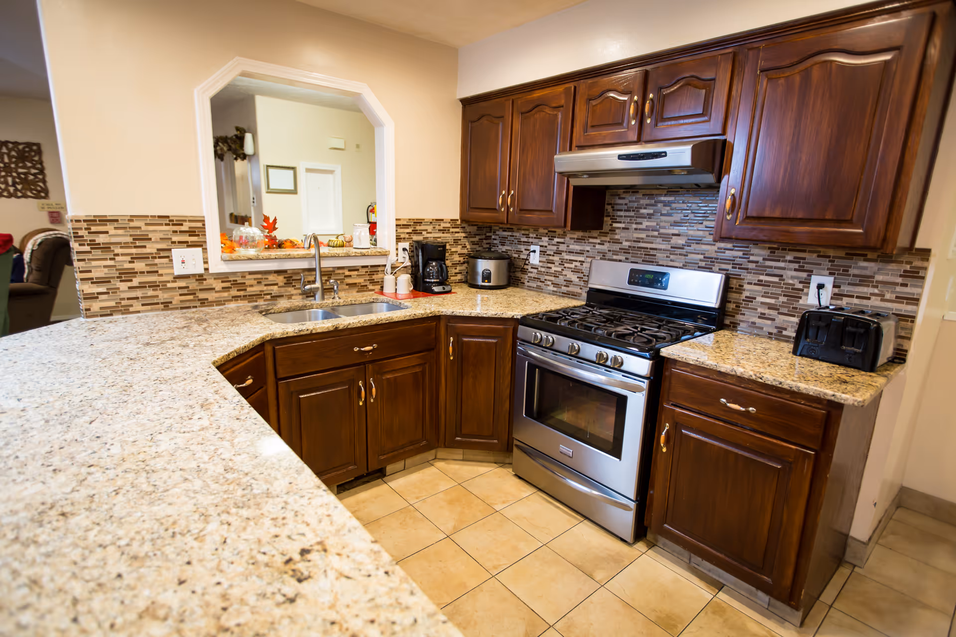 A kitchen area with dark wooden cabinets, a stainless steel gas stove and oven, a toaster, coffee maker, and a rice cooker on granite countertops. The backsplash features a mosaic tile design in neutral tones. There is a double sink under a pass-through window opening to another room.
