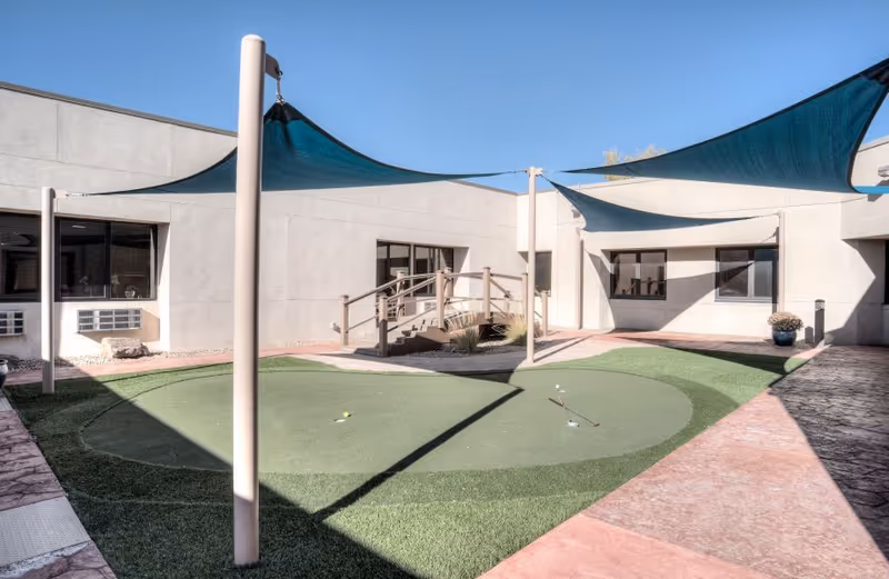 Sunny enclosed courtyard with a small putting green, shade sails, and a wooden footbridge surrounded by the facility's single-story building.