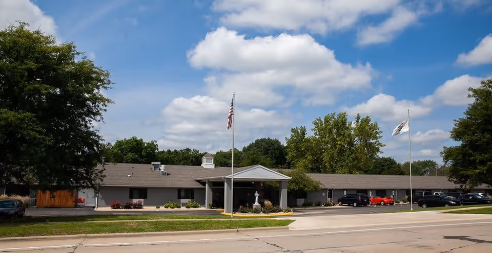 Exterior view of a single-story senior living facility building with a covered entrance, two flagpoles with flags, several parked cars, and trees in the background under a partly cloudy sky.
