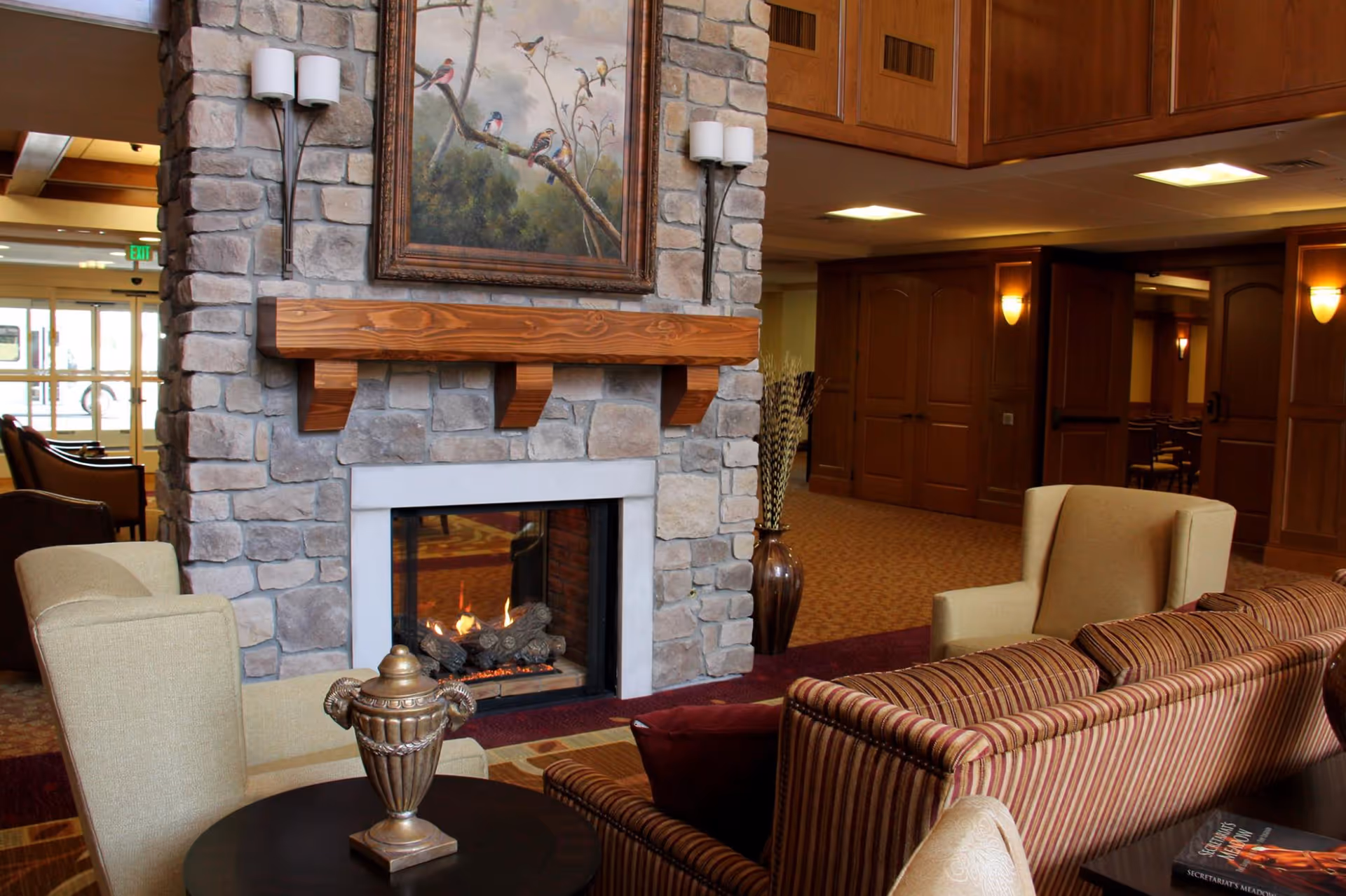 Cozy senior living facility common area with a stone fireplace featuring a wooden mantel and a painting of birds above it. The room includes comfortable beige armchairs, a striped sofa with red and beige cushions, a small round table with a decorative urn, and warm lighting. Wooden doors and carpeted floors are visible in the background.