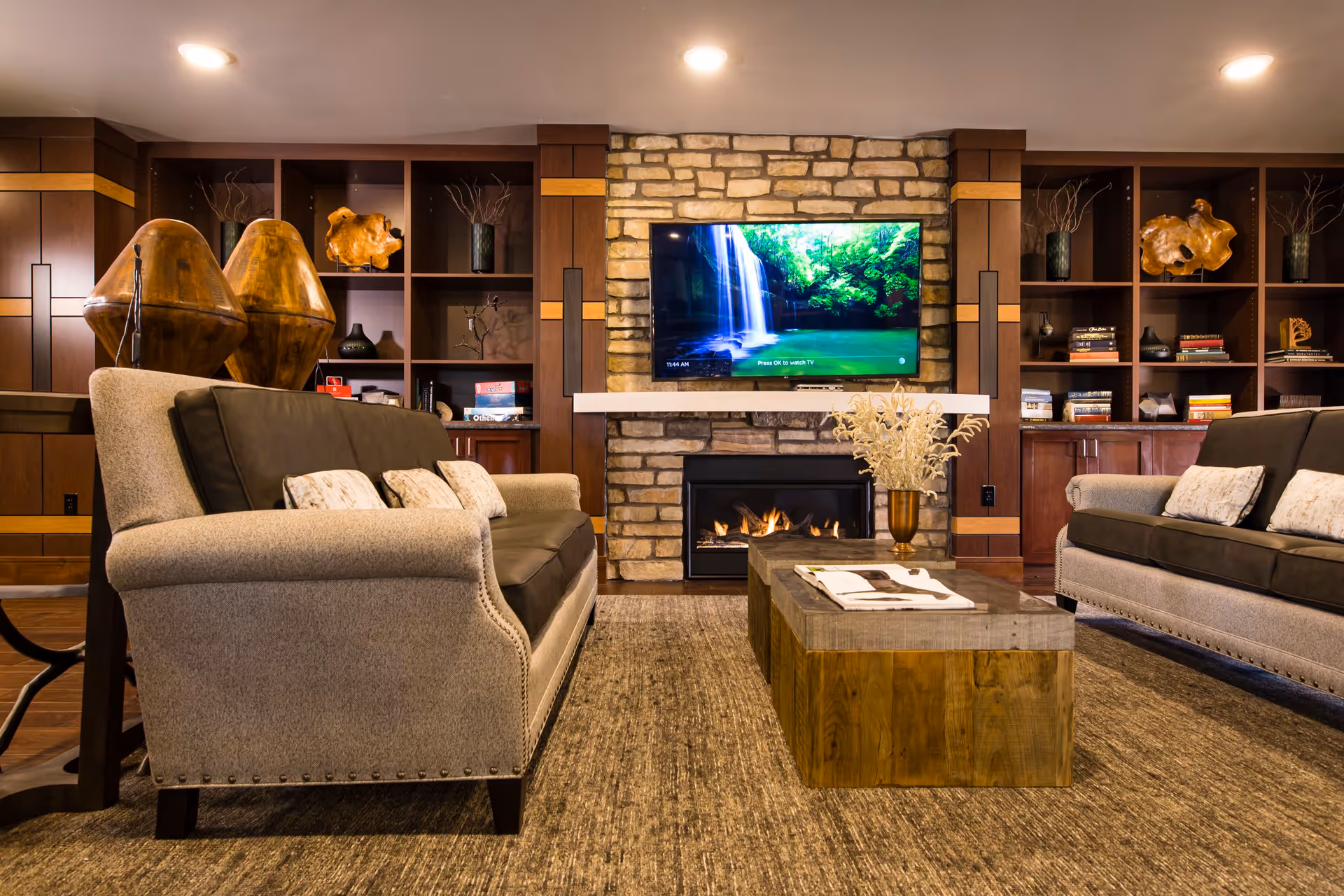 A cozy living room with two beige and brown sofas facing each other, a wooden coffee table with a vase and a magazine, a stone fireplace with a fire burning, and a flat-screen TV mounted above the fireplace displaying a waterfall scene. The background features built-in wooden shelves with decorative items and books.