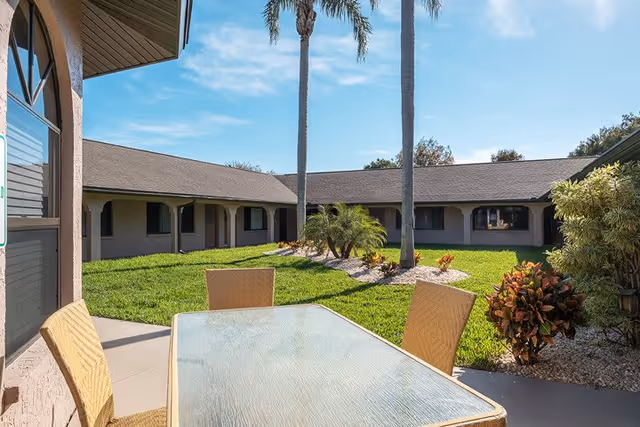 Outdoor courtyard area at Grand Villa of Englewood featuring a glass-top table with four wicker chairs on a concrete patio, surrounded by green grass, palm trees, and various shrubs under a clear blue sky.