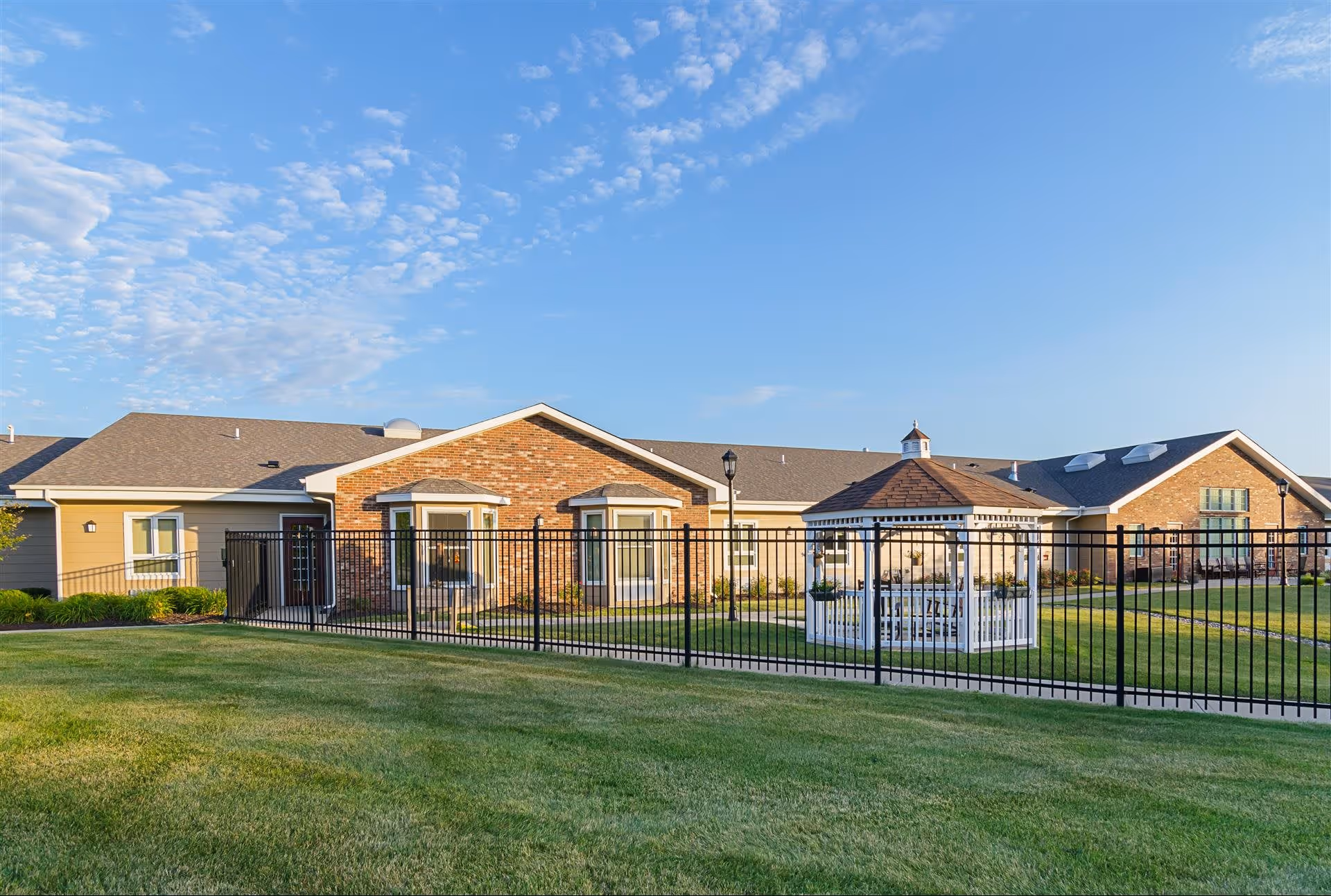 Exterior view of The Gardens at Barry Road Assisted Living and Memory Care facility showing a single-story building with brick and siding, a fenced grassy area, and a white gazebo under a blue sky with scattered clouds.