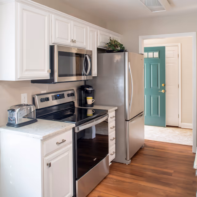 A modern kitchen with white cabinets, a stainless steel refrigerator, stove, and microwave. There is a toaster and a coffee maker on the countertop. The floor is wooden, and a green door is visible in the background.