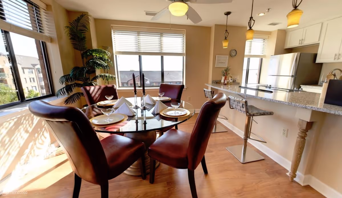 A bright dining area with a round glass table set for four with plates, napkins, and glasses. Four brown leather chairs surround the table. The room features large windows with white blinds, a ceiling fan, a potted plant in the corner, and a kitchen counter with three modern bar stools and pendant lights hanging above.