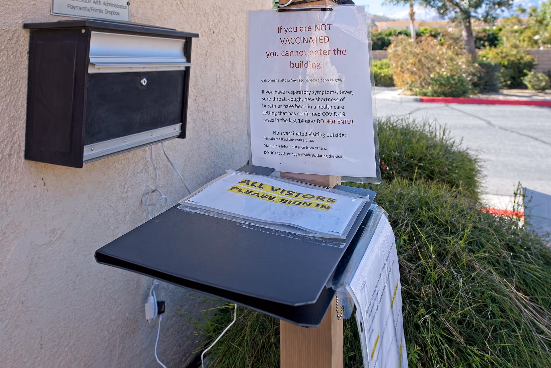 A visitor sign-in station outside a building with a black dropbox mounted on the wall and a clipboard stand holding COVID-19 vaccination and visitor sign-in instructions. There are bushes and a parking lot in the background.