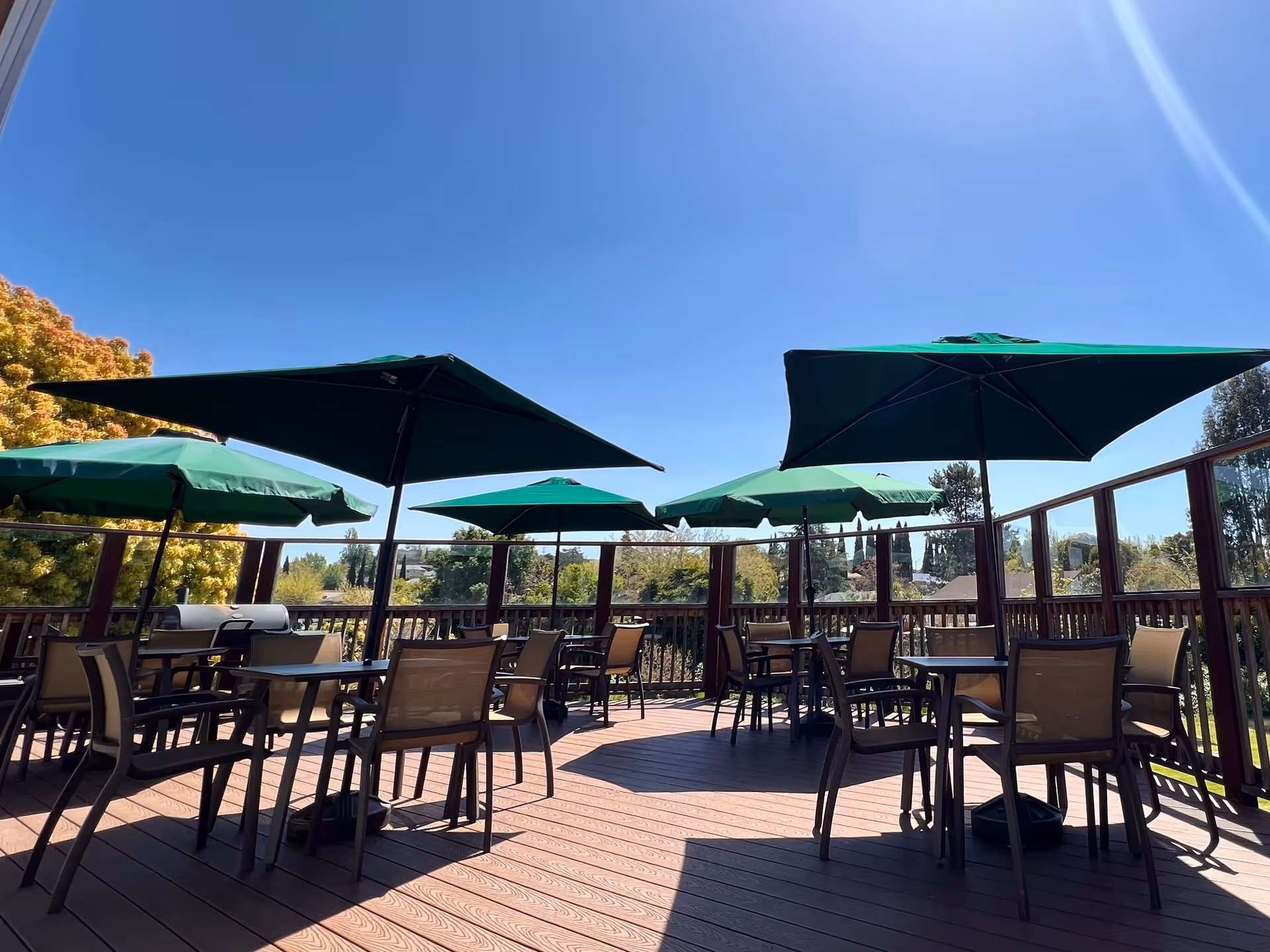 Outdoor patio area with several tables and chairs under green umbrellas on a wooden deck, surrounded by a wooden railing with glass panels, under a clear blue sky.