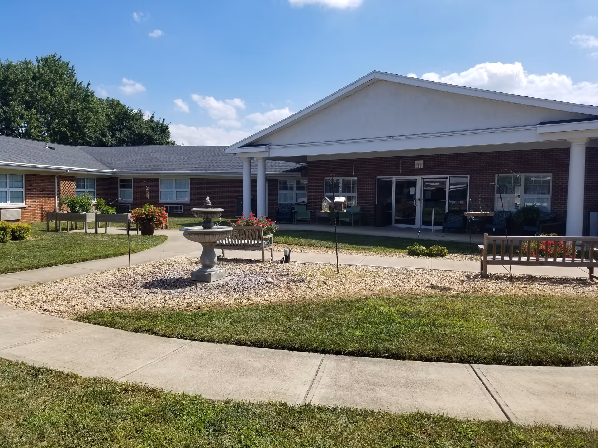 Outdoor courtyard area of The Trinity Community at Fairborn featuring a central stone birdbath surrounded by gravel, two wooden benches, a curved concrete walkway, green grass, raised garden beds, and a brick building with white columns and large windows under a blue sky with scattered clouds.