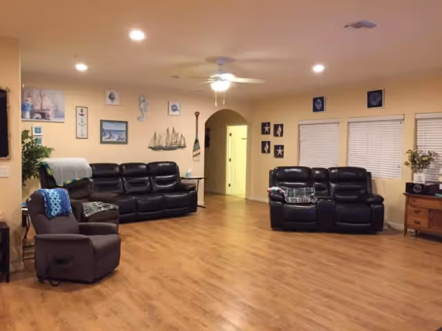 A spacious living room with wooden flooring, beige walls, and a ceiling fan with light. The room features two black leather sofas and one brown recliner chair, each with blankets draped over them. The walls are decorated with nautical-themed artwork including anchors, seahorses, and starfish. There are three windows with white blinds and a wooden side table with a vase of flowers and a radio.