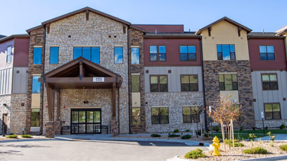 Front exterior view of a multi-story assisted living facility with stone and wood paneling, large windows, and a covered entrance with the number 1111 above it. There is a driveway and landscaped area with small trees and a yellow fire hydrant in front.