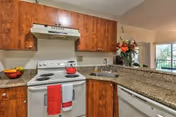 Kitchen area with wooden cabinets, a white electric stove with a red kettle and red towels, granite countertops, a sink, dishwasher, and a bowl of fruit. In the background, there is a living room area with a large window and a floral arrangement on the counter.