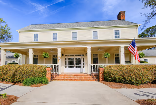Front exterior view of a two-story building with a covered porch supported by columns, hanging plants, an American flag, and neatly trimmed bushes along the entrance pathway under a clear blue sky.
