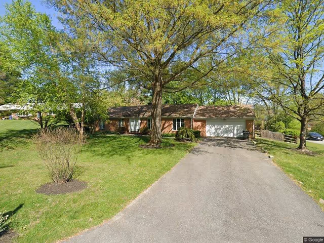 Single-story brick house with a two-car garage, surrounded by green grass and large trees under a clear blue sky.