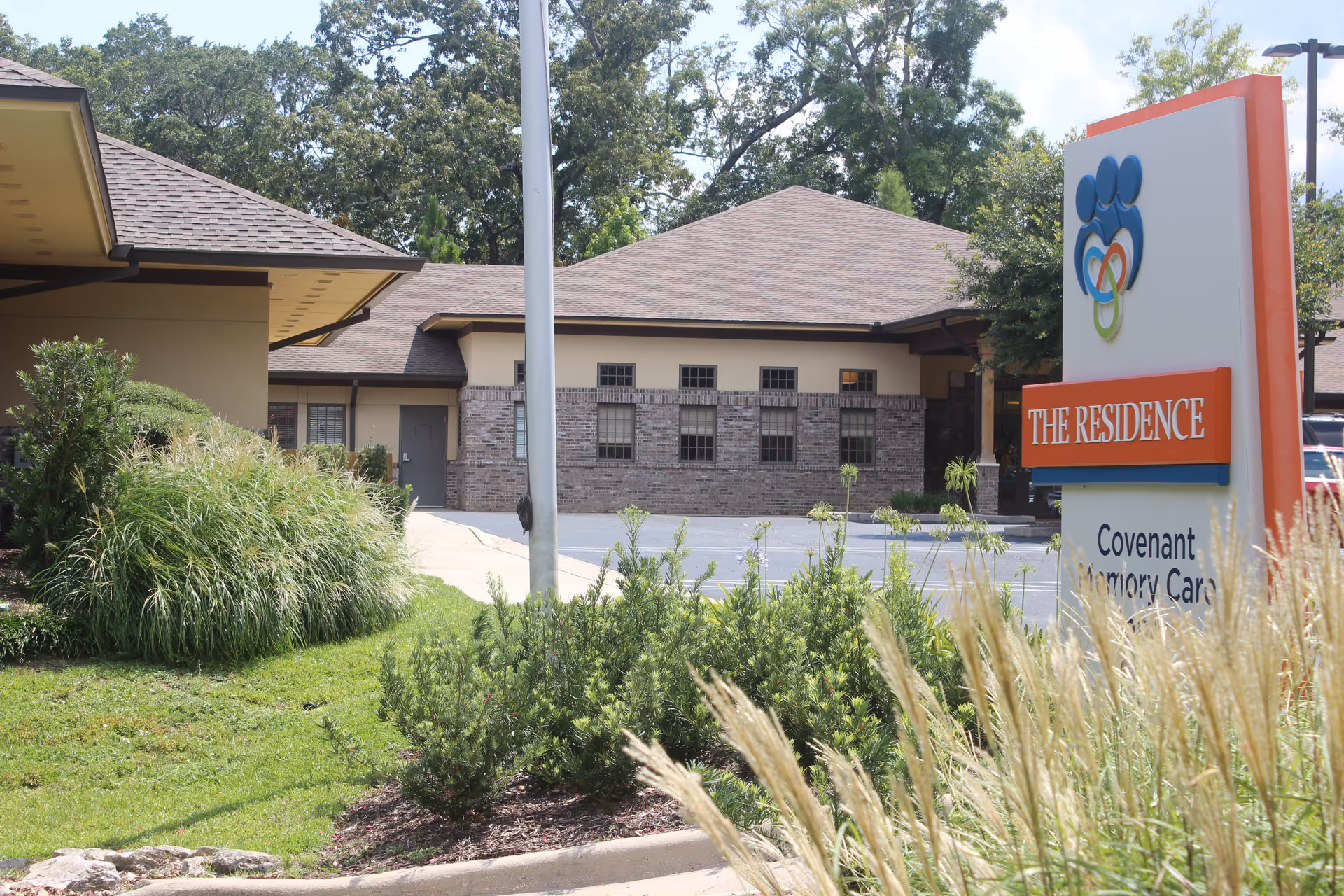 Exterior view of The Residence - VITAS Healthcare facility showing a building with a brick and beige facade, surrounded by greenery and landscaping. A sign in the foreground reads 'THE RESIDENCE Covenant Memory Care' with a colorful logo above it.