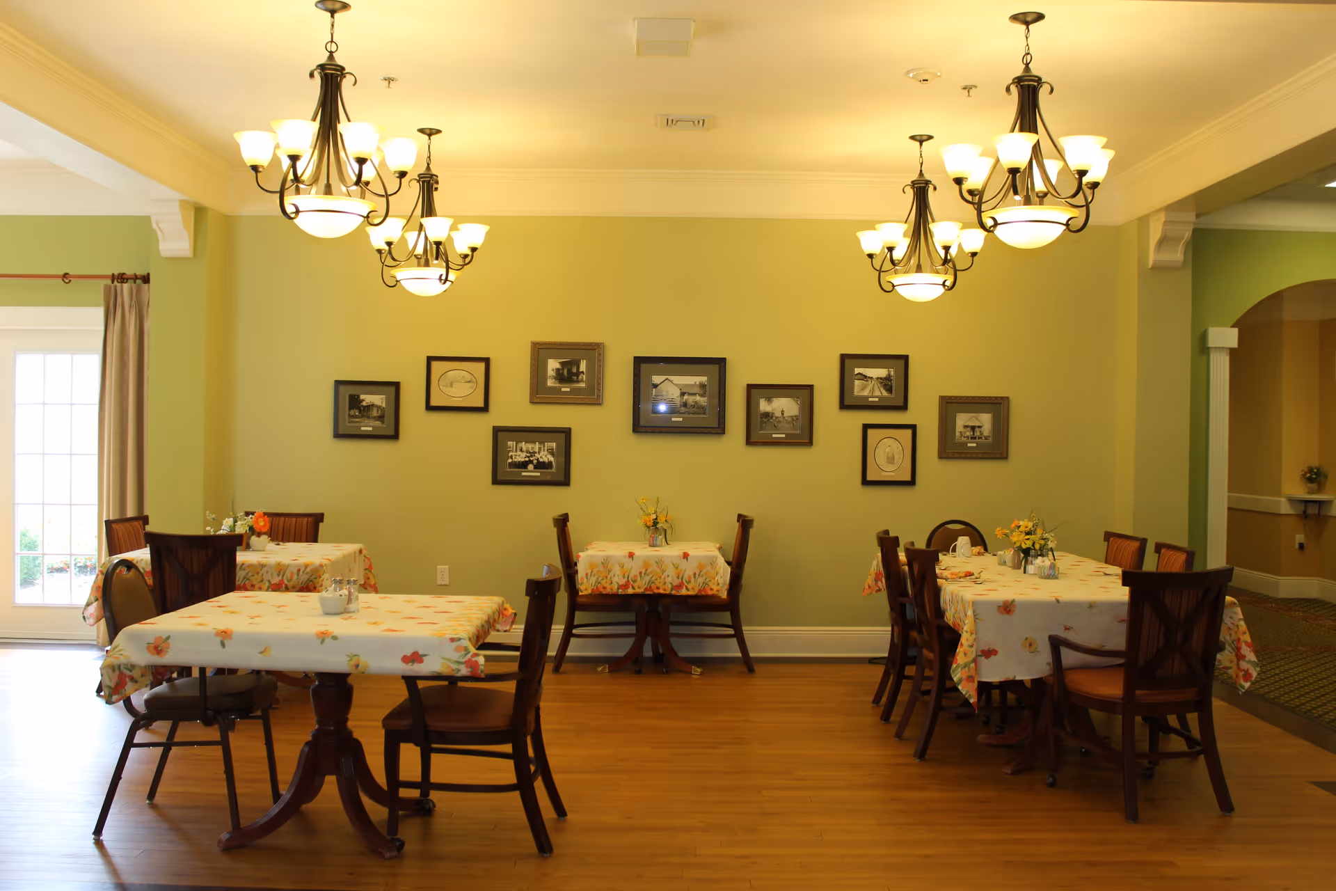 A dining room with several tables covered in floral tablecloths, each surrounded by wooden chairs. The walls are painted light green and decorated with framed black and white photographs. Three chandeliers with multiple lights hang from the ceiling, illuminating the room. A door with glass panels is visible on the left side, and an archway leads to another area on the right.