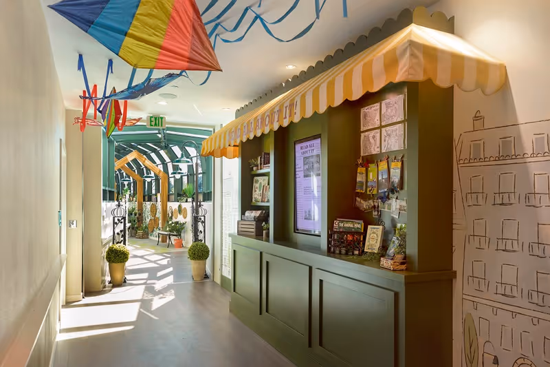 A bright indoor hallway decorated with colorful kites hanging from the ceiling. On the right side, there is a green information kiosk with a yellow and white striped awning and various brochures and notices displayed. The hallway leads to a sunlit area with plants and wooden arch structures.