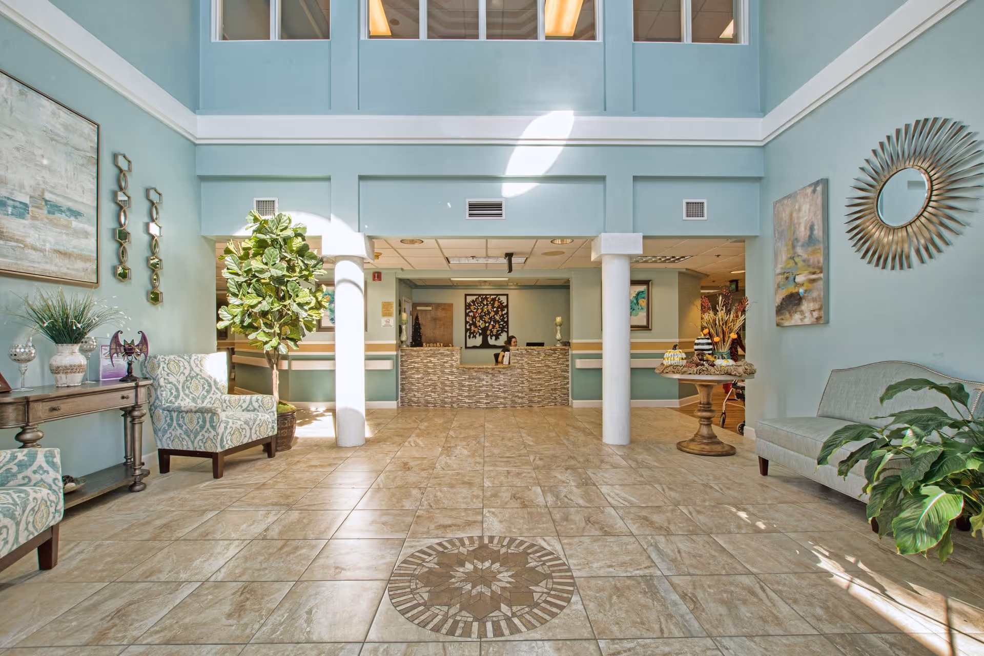 Bright and spacious lobby area of Bridgewater at Waterman Village Assisted Living with tiled floor featuring a decorative circular pattern, light blue walls, and large windows above. The reception desk is straight ahead with a person seated behind it. The room is furnished with patterned armchairs, a small table with decorative items, plants, and wall art including a sunburst mirror and paintings.
