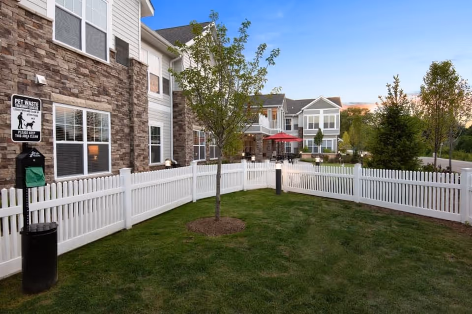 Outdoor area of a senior living facility with a white picket fence enclosing a grassy space with a young tree. The building exterior features stone and siding with multiple windows. There is a pet waste station with a sign and a trash bin. In the background, there are patio tables with red umbrellas and more trees along a paved path.