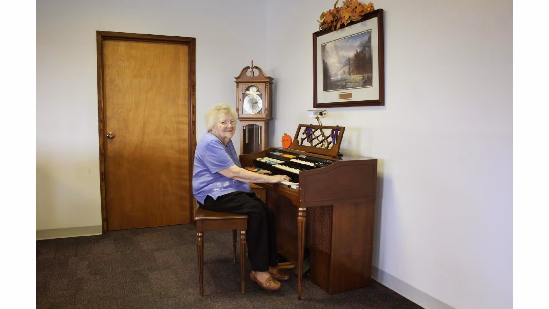 An elderly woman with white hair and glasses, wearing a purple shirt and black pants, is sitting on a wooden chair playing a brown organ in a room with a wooden door, a grandfather clock, and a framed picture on the wall.
