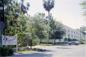 Exterior view of Magnolia Gardens senior living facility with a sign in the foreground, trees, and a parking area with a few cars parked along the driveway.