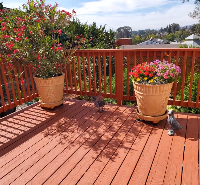 A wooden deck with reddish-brown floorboards and railing. Two large beige flower pots with vibrant flowers and green plants are placed on the deck. Two small decorative bird statues are also on the deck. In the background, there are trees, rooftops, and a partly cloudy sky.