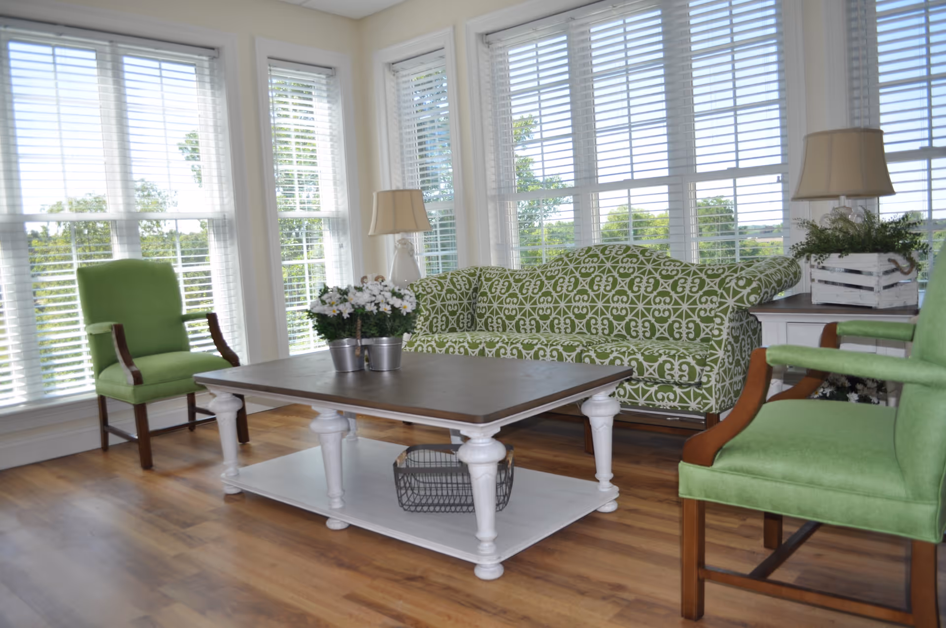 A bright living room with large windows covered by white blinds. The room features a green patterned sofa, two green armchairs with wooden arms, a white coffee table with a dark wooden top, and two table lamps. There are potted plants on the coffee table and a white crate with greenery on a side table.