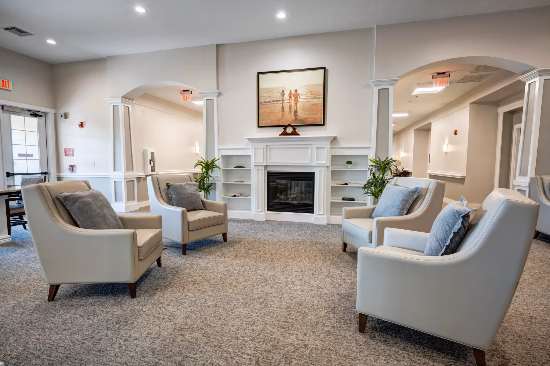 A cozy sitting area in an assisted living facility featuring four beige armchairs with gray cushions arranged around a white fireplace. Above the fireplace is a painting of three children walking on a beach. The room has light-colored walls, carpeted flooring, built-in shelves on either side of the fireplace with decorative items, and two green potted plants. There are arched doorways and exit signs visible in the background.