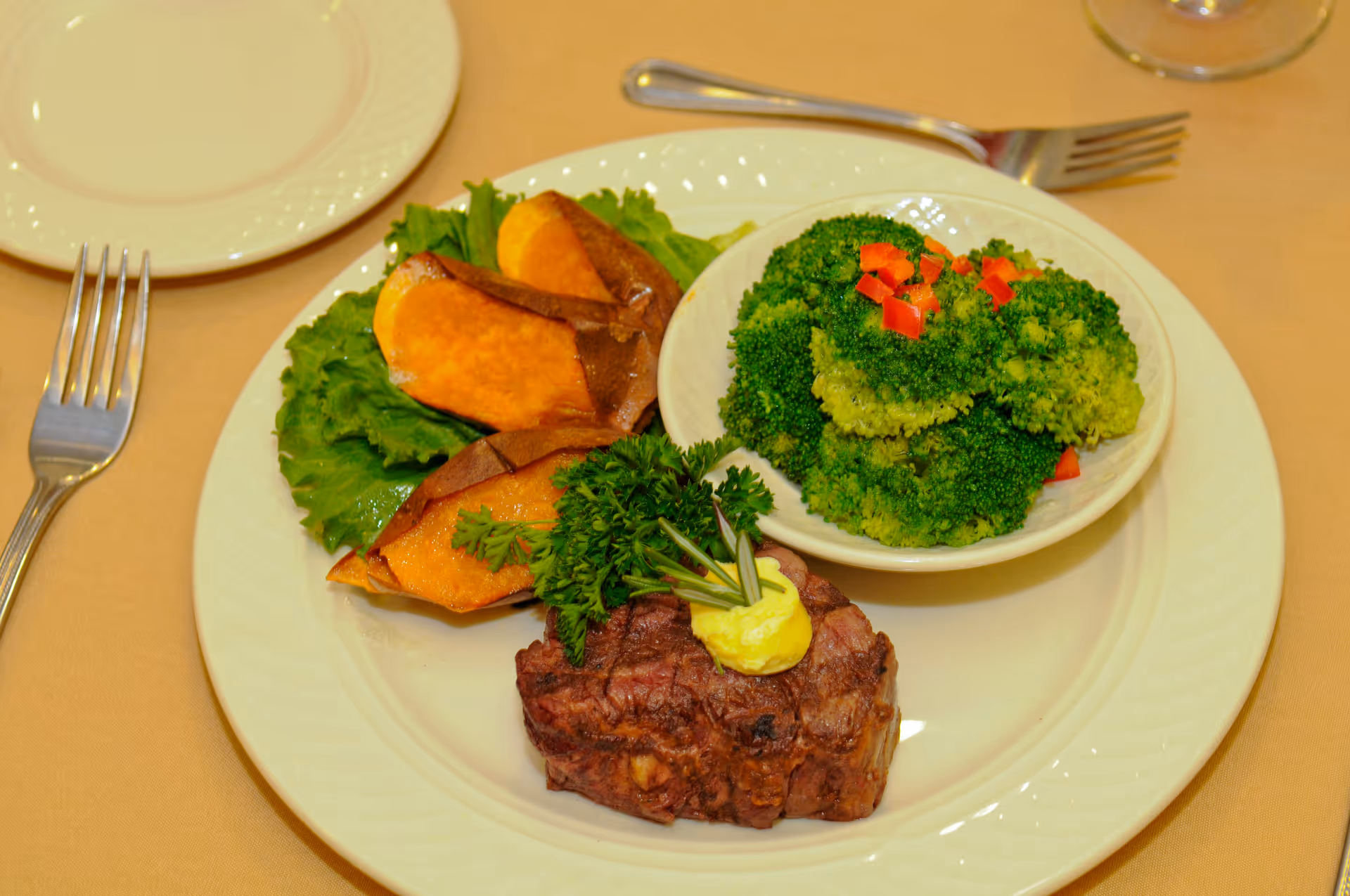 Plate with a grilled steak topped with herb butter and parsley, roasted sweet potato slices, and a small bowl of broccoli on a dining table.
