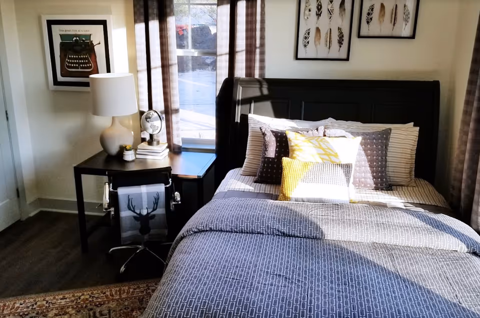 Sunlit bedroom with a made bed, patterned bedding and pillows, a nightstand with a lamp and globe, and a window with curtains.