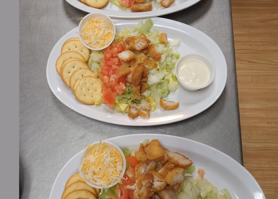 Two white oval plates each containing a salad with chopped lettuce, diced tomatoes, fried chicken pieces, a small cup of shredded cheese, several round crackers, and a small cup of creamy dressing on a stainless steel surface next to a wooden floor.