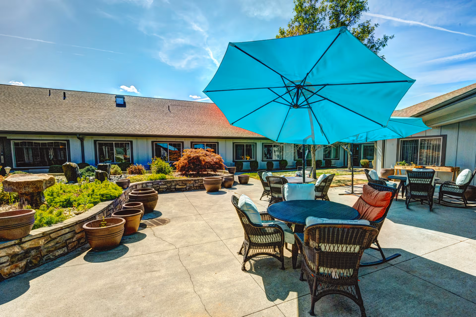 Outdoor patio area at Pine Ridge Alzheimer's Special Care with round tables and wicker chairs, some with cushions, shaded by large blue umbrellas. The patio is surrounded by a low stone wall with potted plants and greenery, and the building with multiple windows is visible in the background under a clear blue sky.