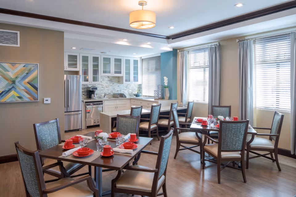 A bright dining area with two wooden tables set with red cups, plates, and white napkins. The room features large windows with blinds and curtains, light wood flooring, and a modern kitchen in the background with white cabinets, stainless steel appliances, and a decorative backsplash. A colorful abstract painting hangs on the wall to the left.