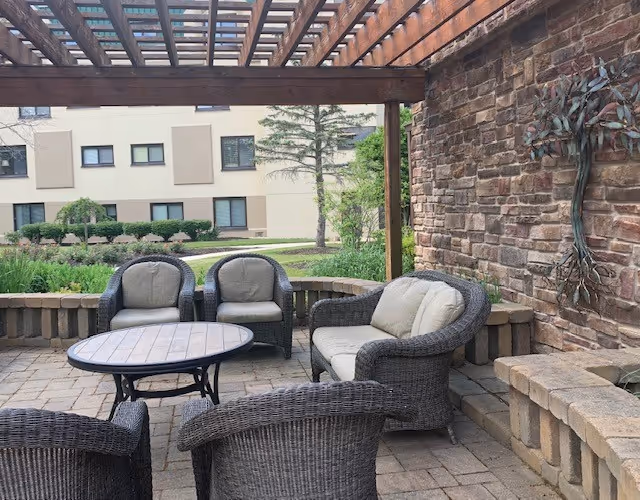 Outdoor patio area with wicker furniture including a round table, two armchairs, a loveseat, and two additional chairs under a wooden pergola. The patio is paved with stone tiles and has a stone wall with a metal tree decoration. In the background, there is greenery and a beige building with windows.