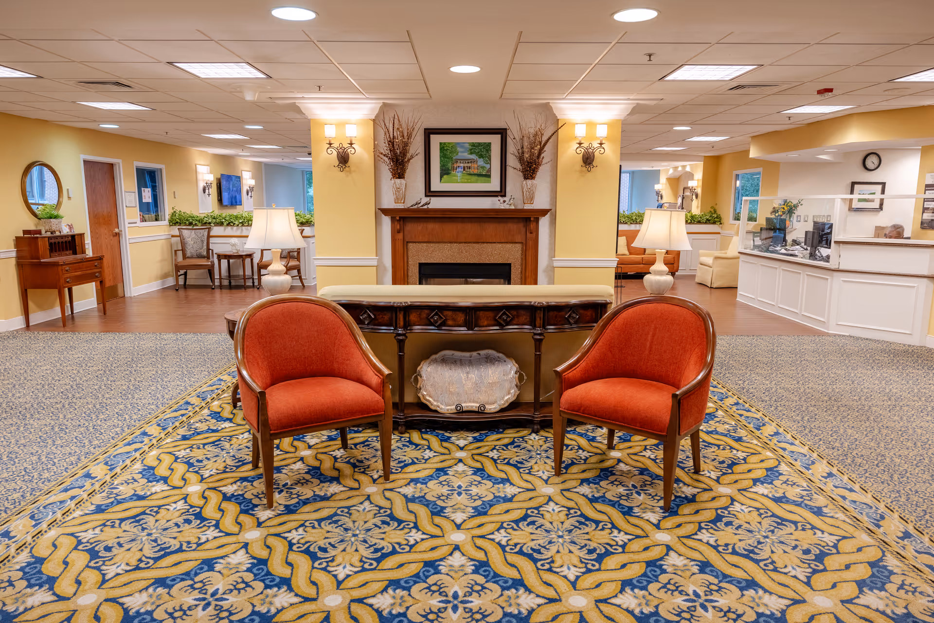 A cozy and well-lit common area in a senior living facility featuring two red upholstered chairs on a patterned blue and gold rug, a wooden console table with a decorative silver tray, and a fireplace with a framed painting above it. The walls are painted yellow with white trim, and there are lamps and wall sconces providing warm lighting. In the background, there is a reception desk with a protective glass barrier and seating areas with chairs and tables.