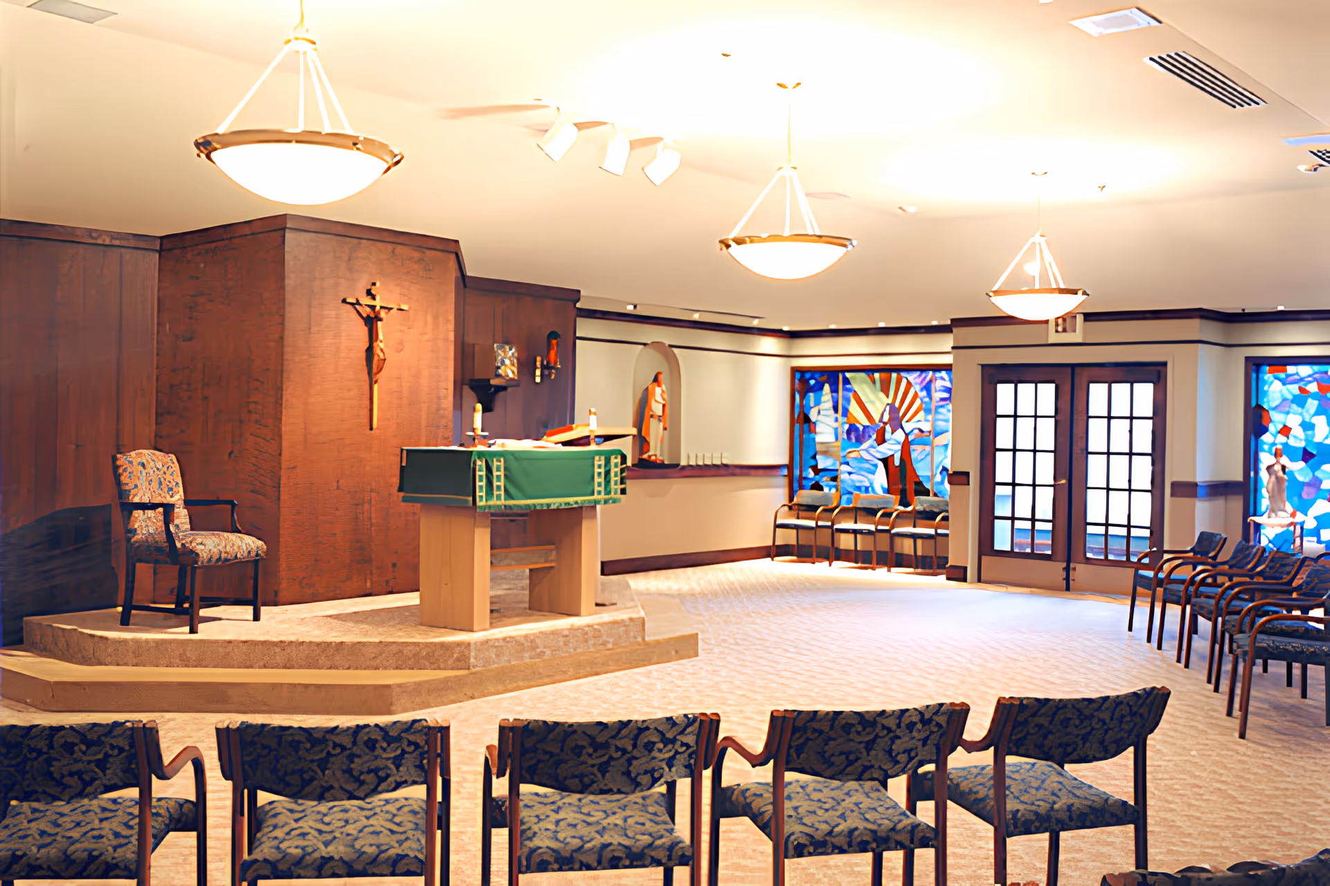 Interior view of a chapel or worship room with a raised platform featuring a wooden crucifix on the wall, a lectern draped with a green cloth, and a single upholstered chair. The room has rows of patterned chairs facing the platform, stained glass windows depicting religious scenes, and soft overhead lighting.