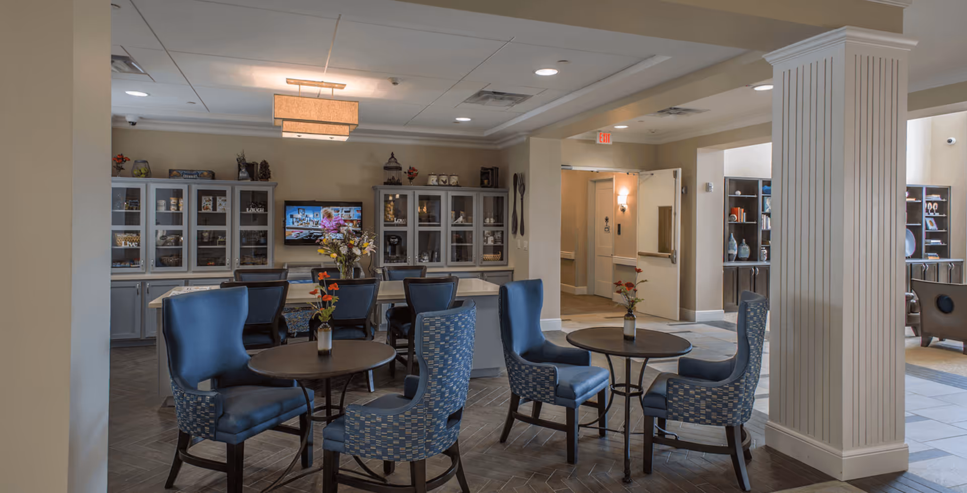 A cozy common area in a senior living facility featuring several round tables with blue upholstered chairs. Each table has a small vase with flowers. In the background, there are gray cabinets with glass doors displaying decorative items, and a flat-screen TV mounted on the wall. The room is well-lit with ceiling lights and a hanging light fixture, and there is an open doorway leading to another area of the facility.