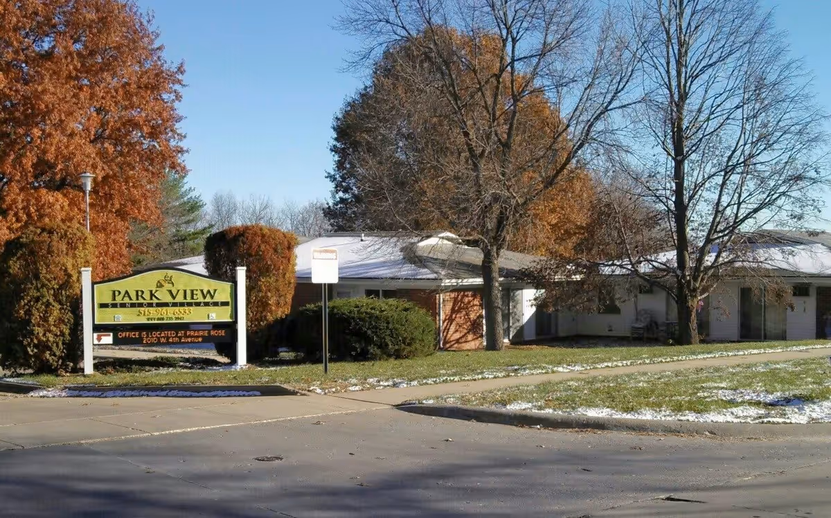 Front exterior of Park View Senior Village showing the low building, trees, lawn with a light dusting of snow, and a prominent Park View sign by the street.