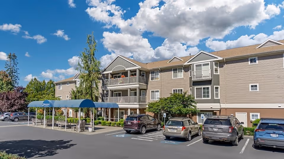Exterior view of a three-story senior living facility building under a blue sky with scattered clouds. Several cars are parked in front of the building, which features balconies and a covered entrance with a blue awning labeled 'Brookdale Olympia East'. Trees and shrubs surround the parking area.