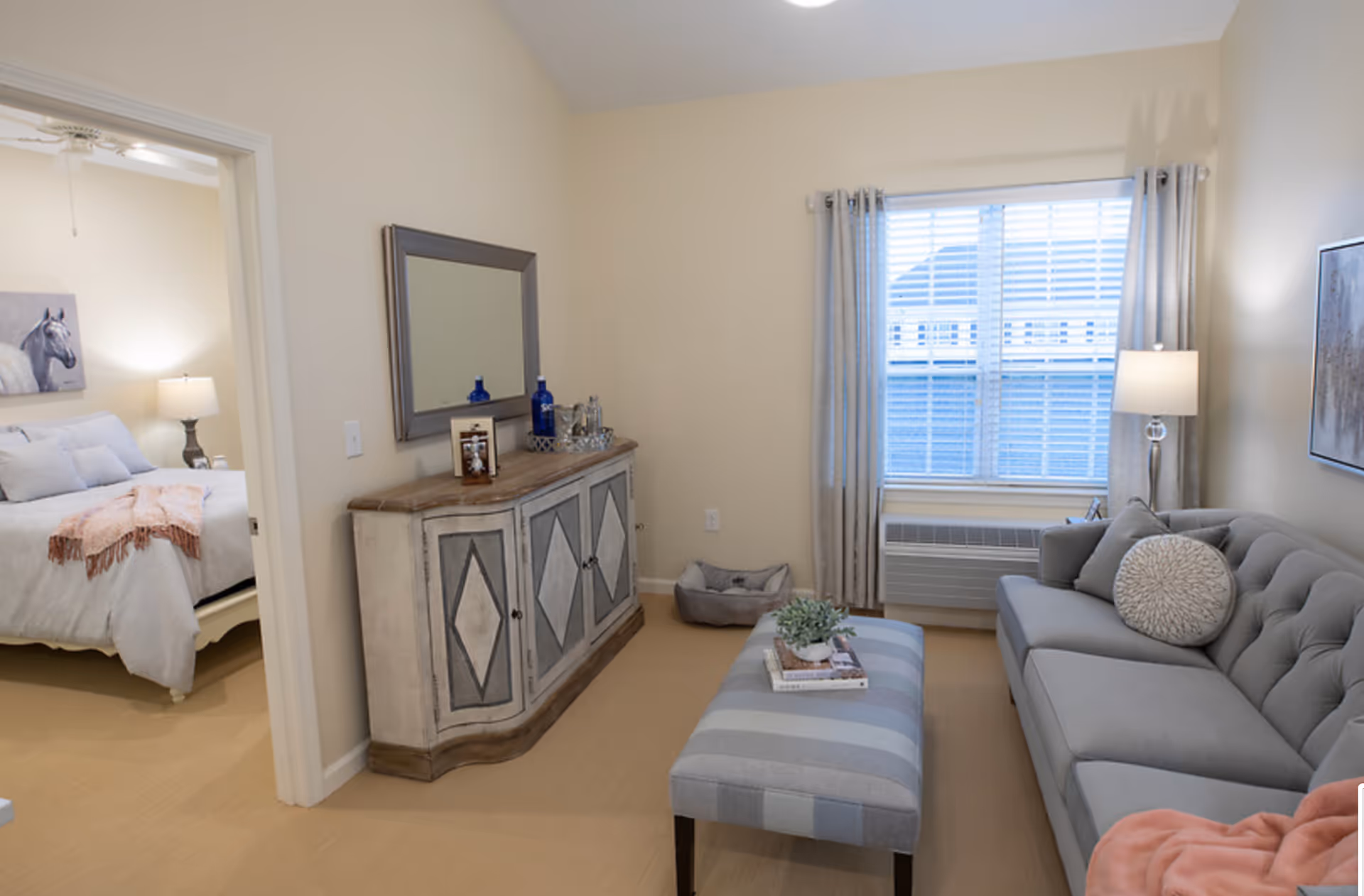 Bright living room with a gray tufted sofa, striped ottoman, sideboard with mirror, and a view into an adjacent bedroom.