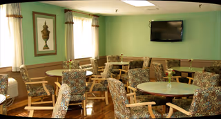 A shared dining lounge with round tables, floral chairs, and a wall-mounted TV.