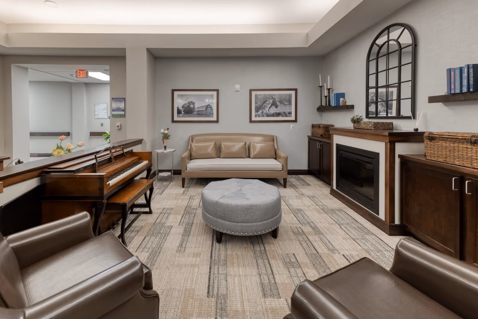 A cozy assisted living facility common area with a beige sofa, two brown leather armchairs, a round gray ottoman, a wooden piano, and a fireplace with a mirror above it. The room has neutral-colored walls and carpet, with framed black and white photos on the wall and decorative items on shelves.