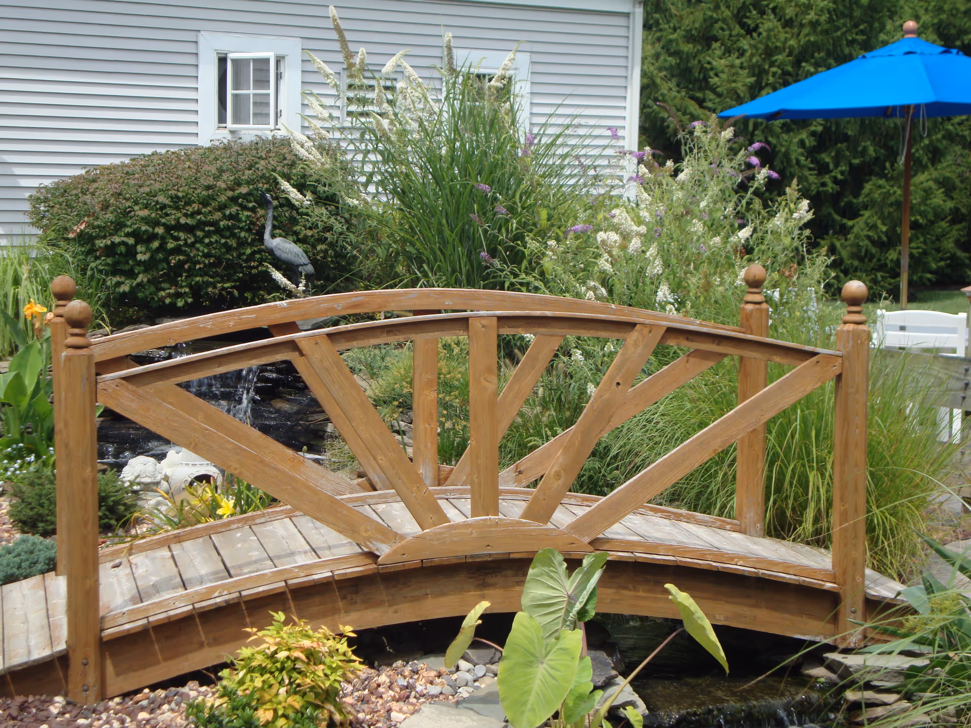 Wooden decorative footbridge over a small garden pond surrounded by plants and a blue patio umbrella in the background.