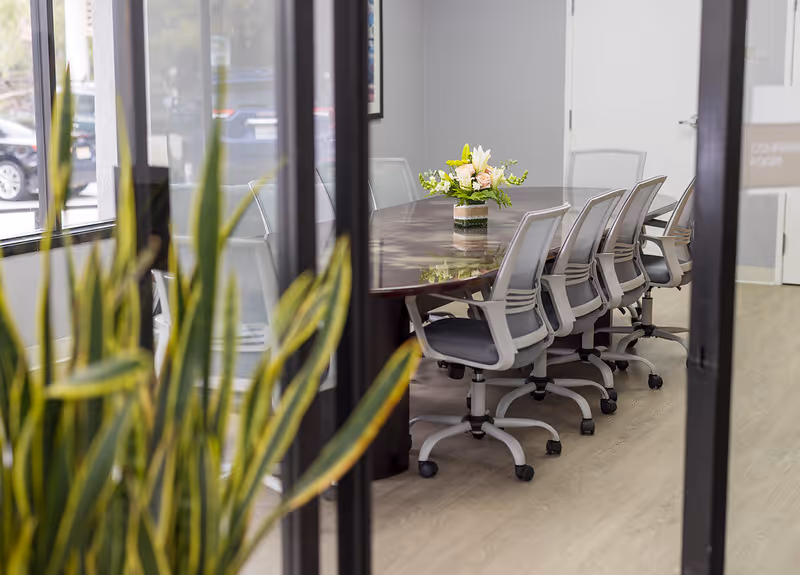 View through glass doors of a conference room with a long wooden table, rolling chairs, and a floral centerpiece.